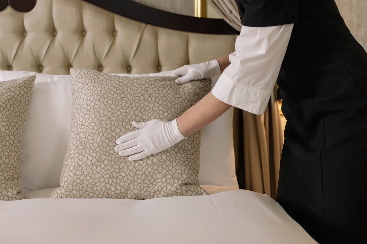 Home A housekeeper wearing gloves arranges pillows on a luxurious bed in a hotel room.